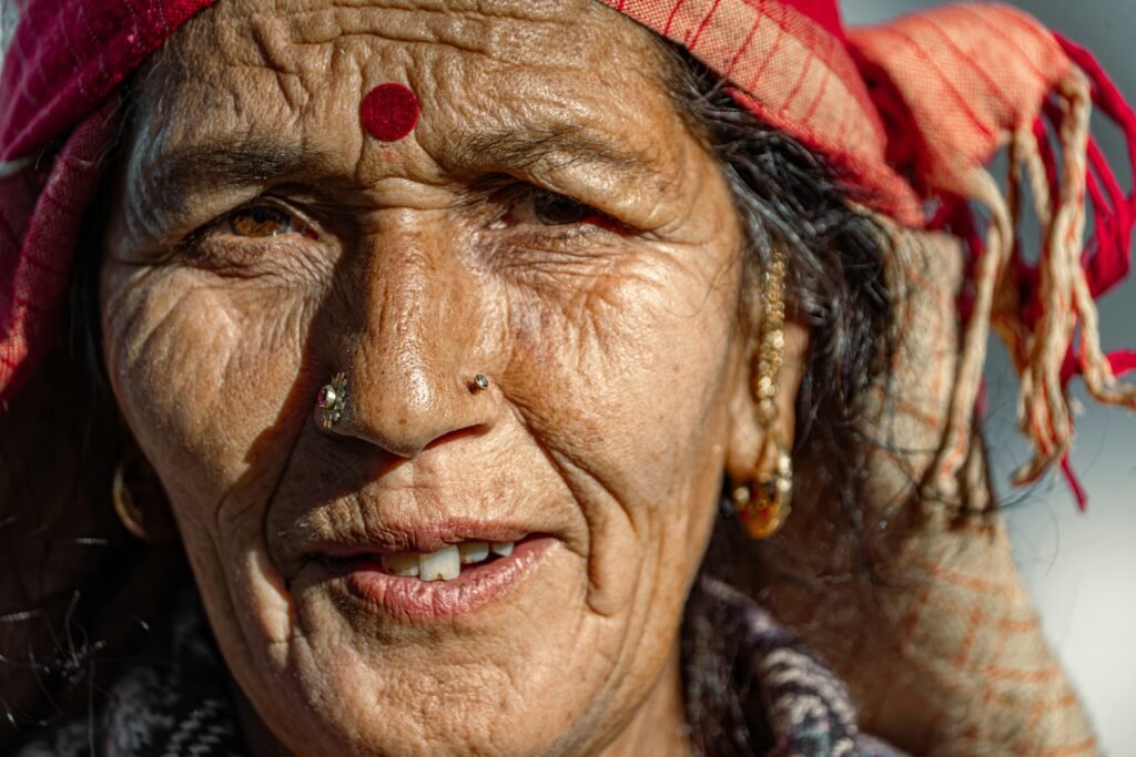 Close-up portrait of an older North Indian woman in a red headscarf and gold nose ring, smiling in natural light